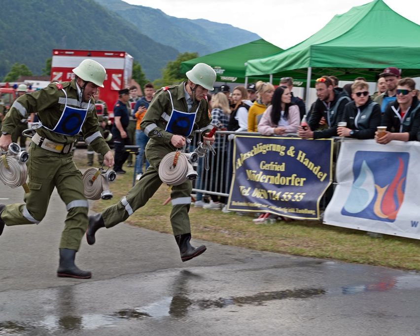 Zwei Feuerwehrleute in grünen Uniformen laufen mit einem Schlauch auf einem nassen Bereich. Zuschauer schauen hinter einem Zaun mit Bannern zu. Berge und ein rotes Rettungsfahrzeug sind im Hintergrund.