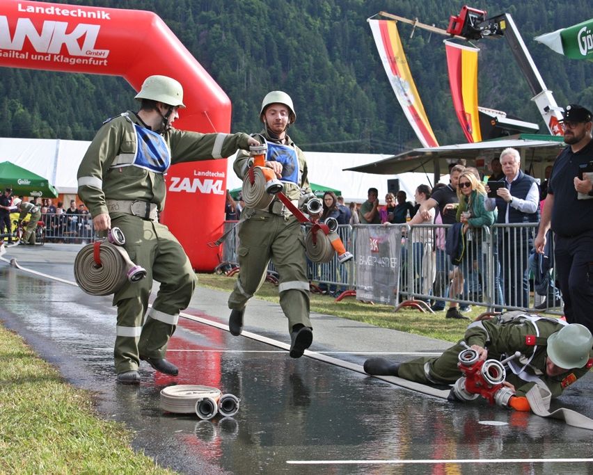Zwei Feuerwehrleute laufen in einem Wettbewerb mit Schläuchen, ein Dritter liegt am Boden. Zuschauer beobachten hinter einem Zaun. Dahinter steht ein roter Bogen mit der Aufschrift 'ZANKL GmbH'.