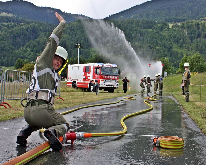 Ein Feuerwehrmann in Uniform signalisiert während einer Feuerwehrübung mit einem Wasserwagen, der Wasser sprüht. In der Nähe handhaben andere Feuerwehrleute Schläuche auf einer nassen Straße.