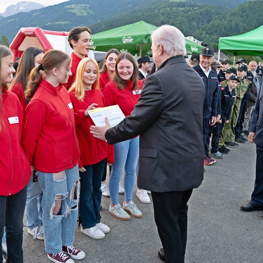 Ein Mann in einem Anzug überreicht einer Frau in einem roten Shirt ein Papier vor einem grünen Zelt. Dahinter stehen weitere Personen in roten Shirts und Kinder in Uniformen. Im Hintergrund sind Berge zu sehen.