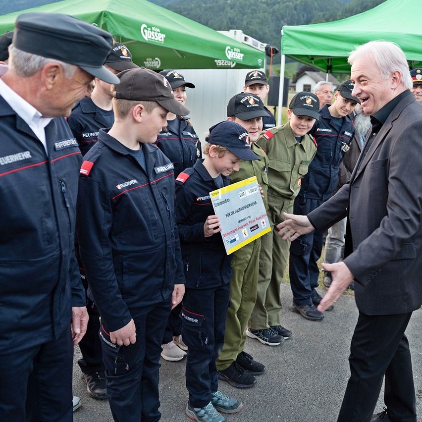 Eine Gruppe von Kindern in Uniformen steht mit einem Mann, der einen Spendenscheck in der Hand hält. Sie sind um grüne Zelte mit Logos versammelt. Der Mann lächelt und unterhält sich mit den Kindern.