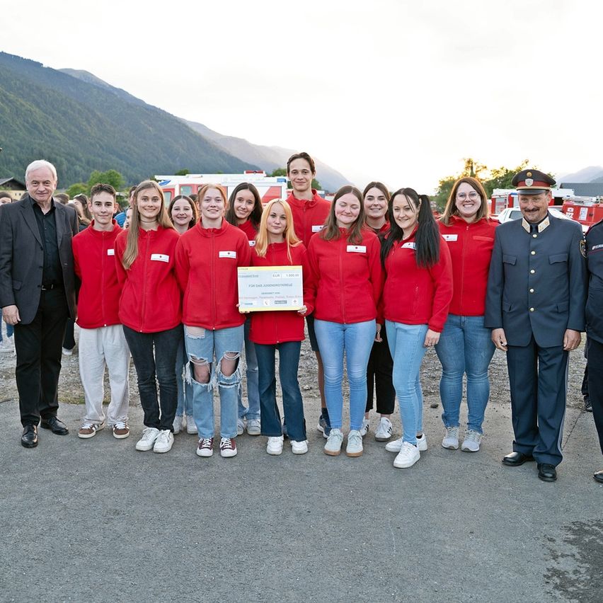 Eine Gruppe junger Menschen in roten Jacken posiert für ein Foto mit einem Mann in Uniform, der ein Zertifikat hält, mit Bergen im Hintergrund.