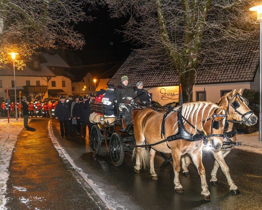 Eine Pferdekutsche fährt bei Nacht eine verschneite Straße hinunter, wobei Menschen in Mänteln am Straßenrand stehen. Die Kutsche wird von zwei Pferden gezogen, und im Hintergrund befindet sich ein Gebäude.
