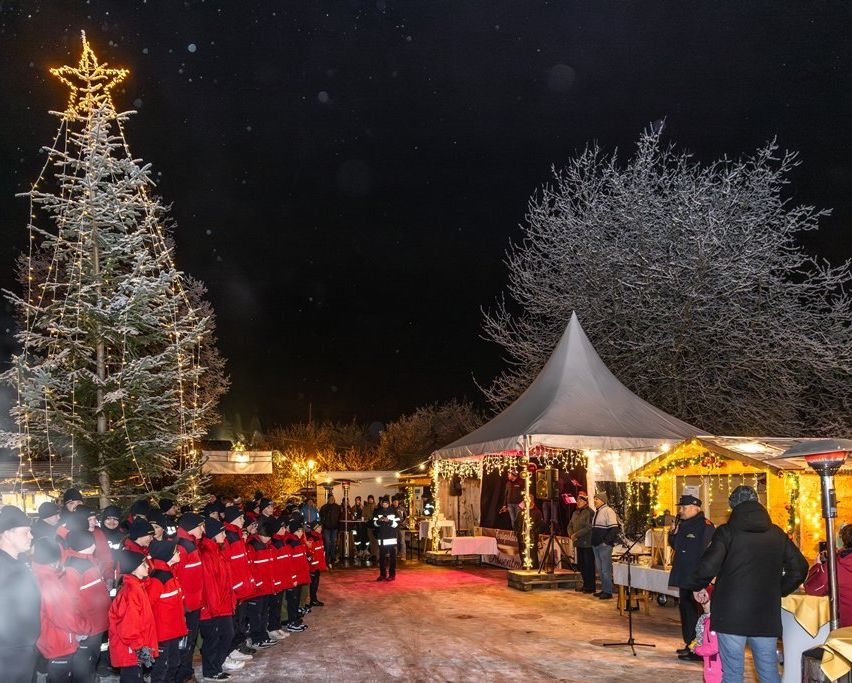 Ein Chor in roten Jacken singt unter einem beleuchteten Weihnachtsbaum und einem Zelt mit festlichen Lichtern und Dekorationen. Mehrere Leute stehen herum, darunter ein Mann, der an einem Mikrofon spricht.