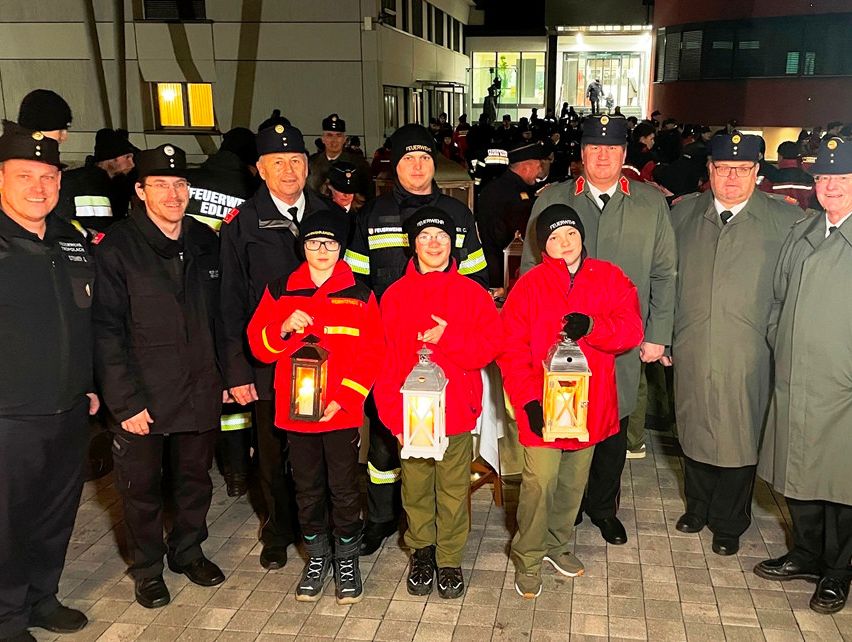 Eine Gruppe von Feuerwehrleuten, sowohl Erwachsene als auch Jugendliche, steht vor einem Gebäude und hält Laternen. Sie sind von anderen Leuten in Uniformen umgeben.