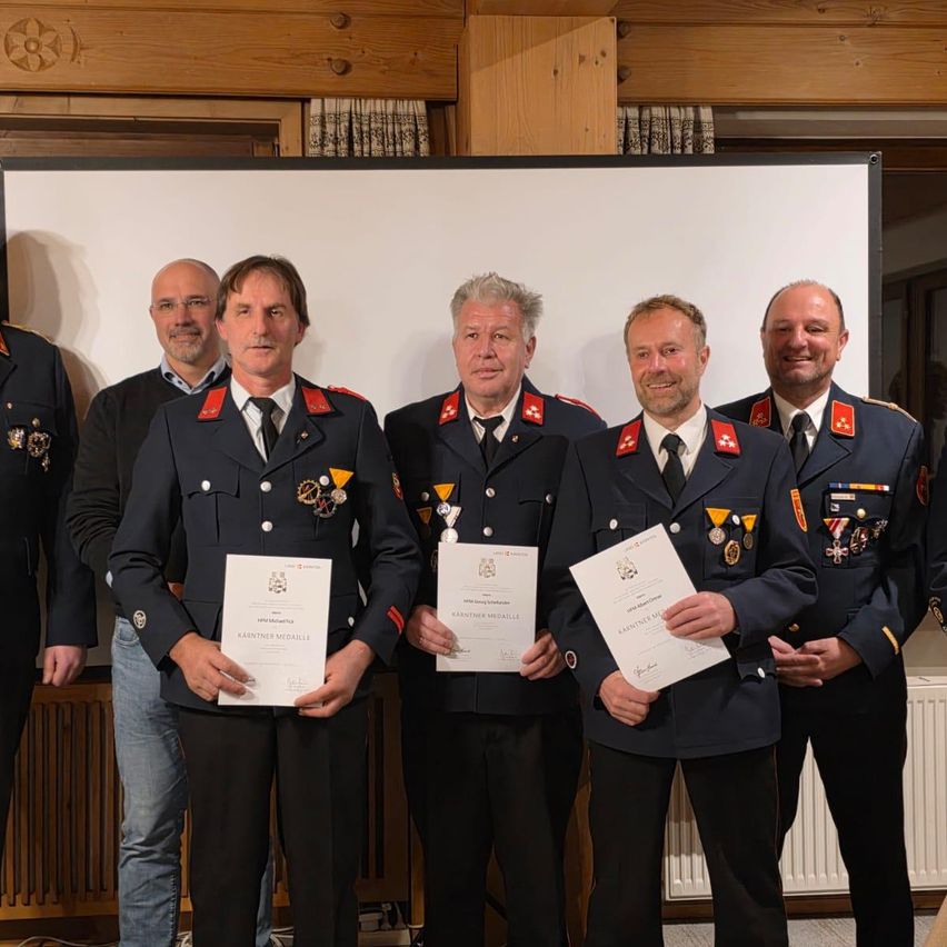 A group of men in uniforms stand together in front of a white backdrop, holding certificates. They are smiling and appear to be celebrating an achievement.