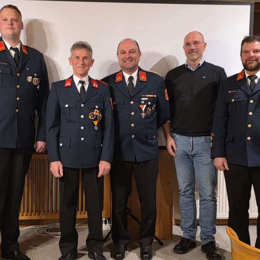Five men stand together in a room, wearing uniforms. They are smiling and seem to be posing for a photograph. Behind them, there is a white board and a radiator.