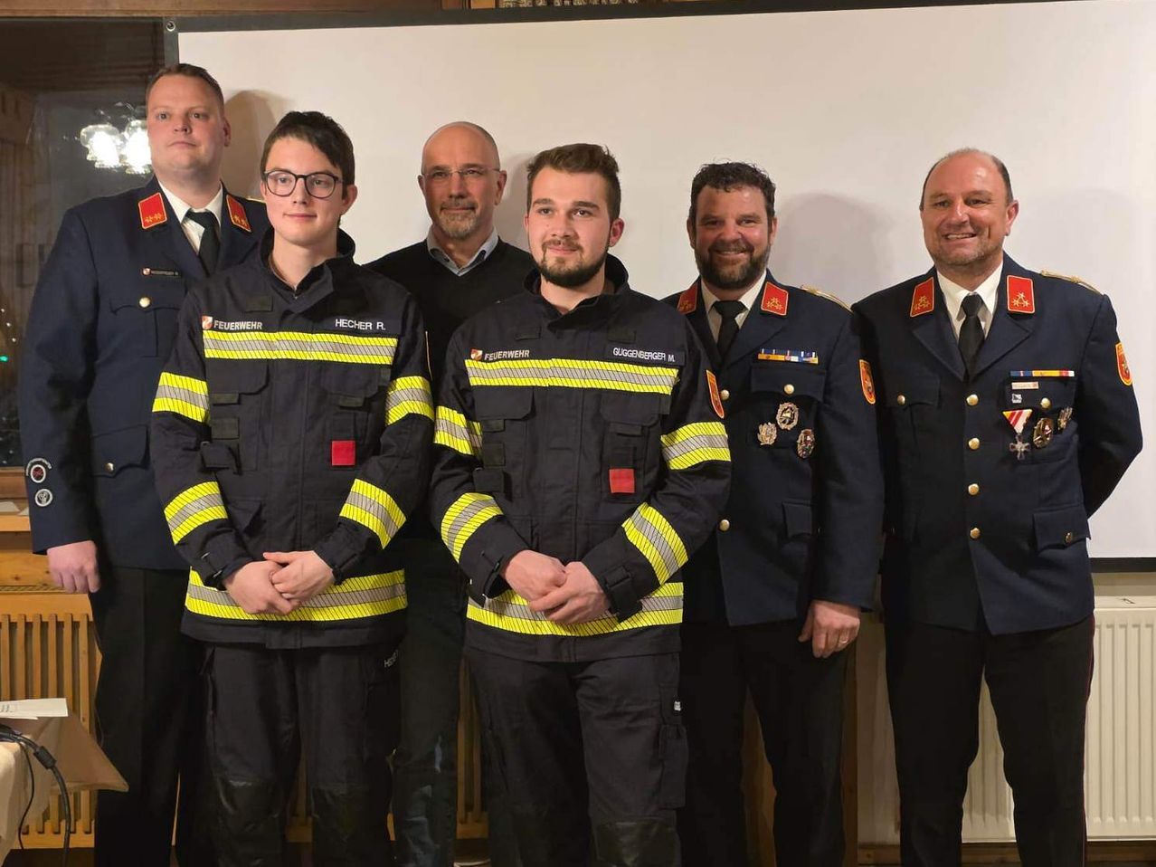 A group of firefighters in uniform pose for a photo. They are standing in front of a white wall, smiling and looking at the camera.