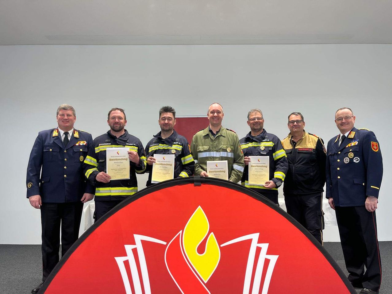 Sieben Feuerwehrleute stehen vor einem roten Banner mit Flammenlogo und halten Zertifikate in den Händen. Sie tragen alle Uniformen, einige mit gelb-schwarzen Streifen.