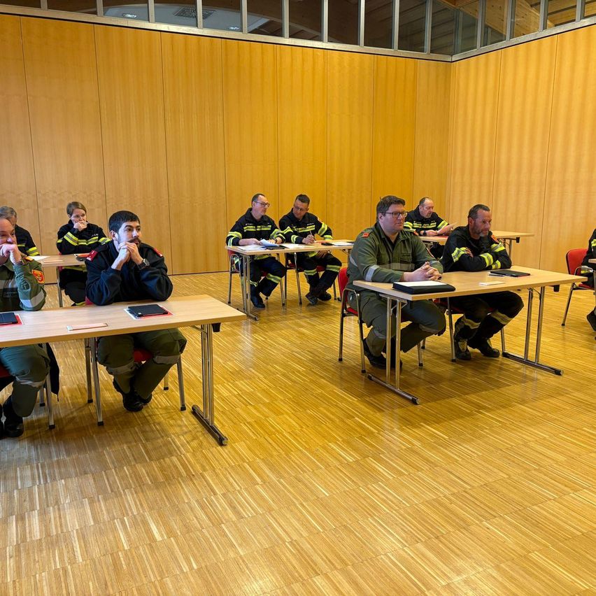 Mehrere Feuerwehrleute in Uniform sitzen an einem Tisch in einem Schulungsraum. Sie scheinen an einer Besprechung teilzunehmen, wobei Papiere und Notizbücher auf dem Tisch liegen. Der Raum hat Holzwände und einen Holzboden.