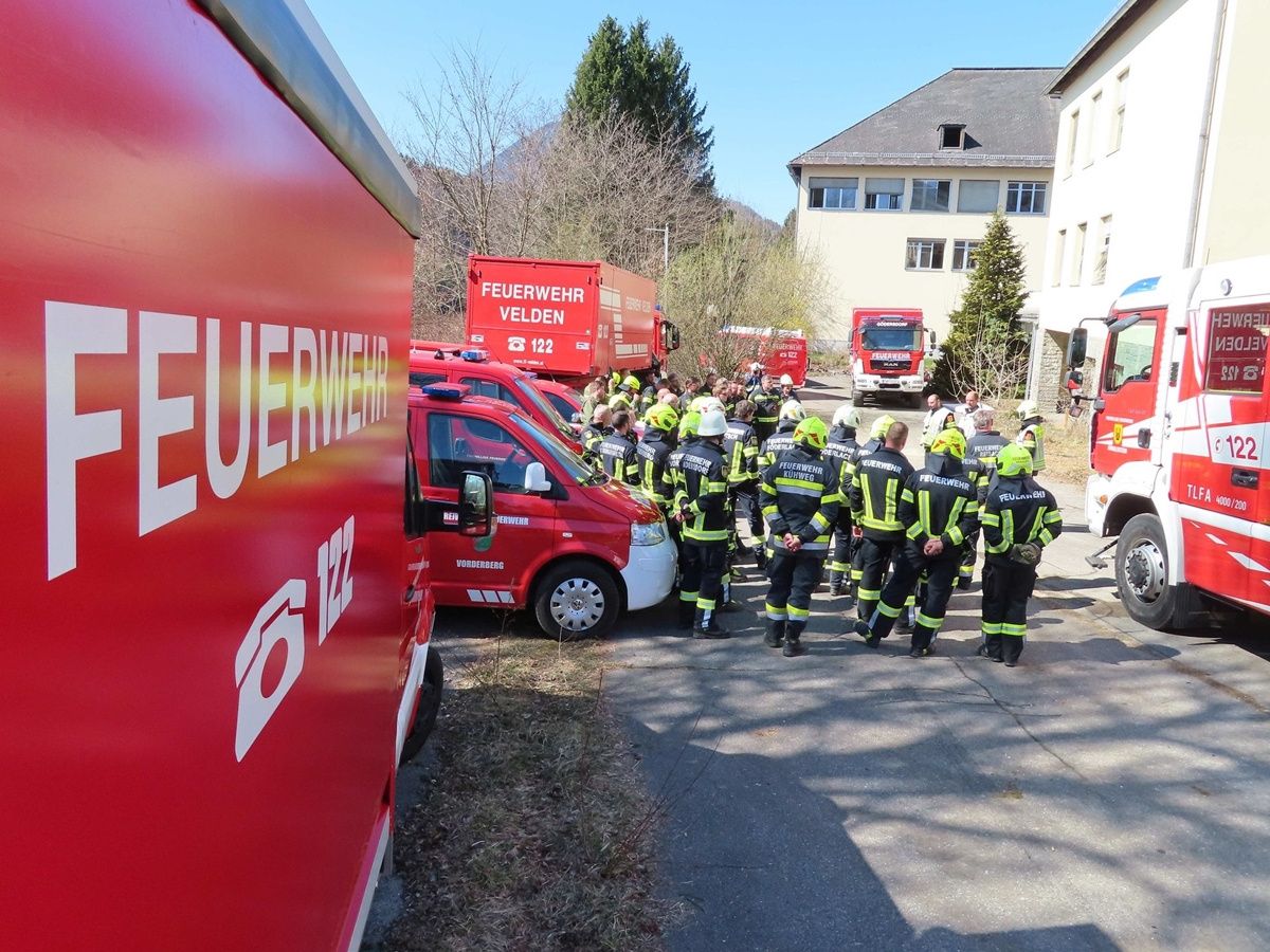 Feuerwehrleute in Uniform versammeln sich vor Gebäuden, mit geparkten Feuerwehr- und Rettungsfahrzeugen. Die Szene spielt sich im Freien ab, mit klarem Himmel und einigen Bäumen im Hintergrund.