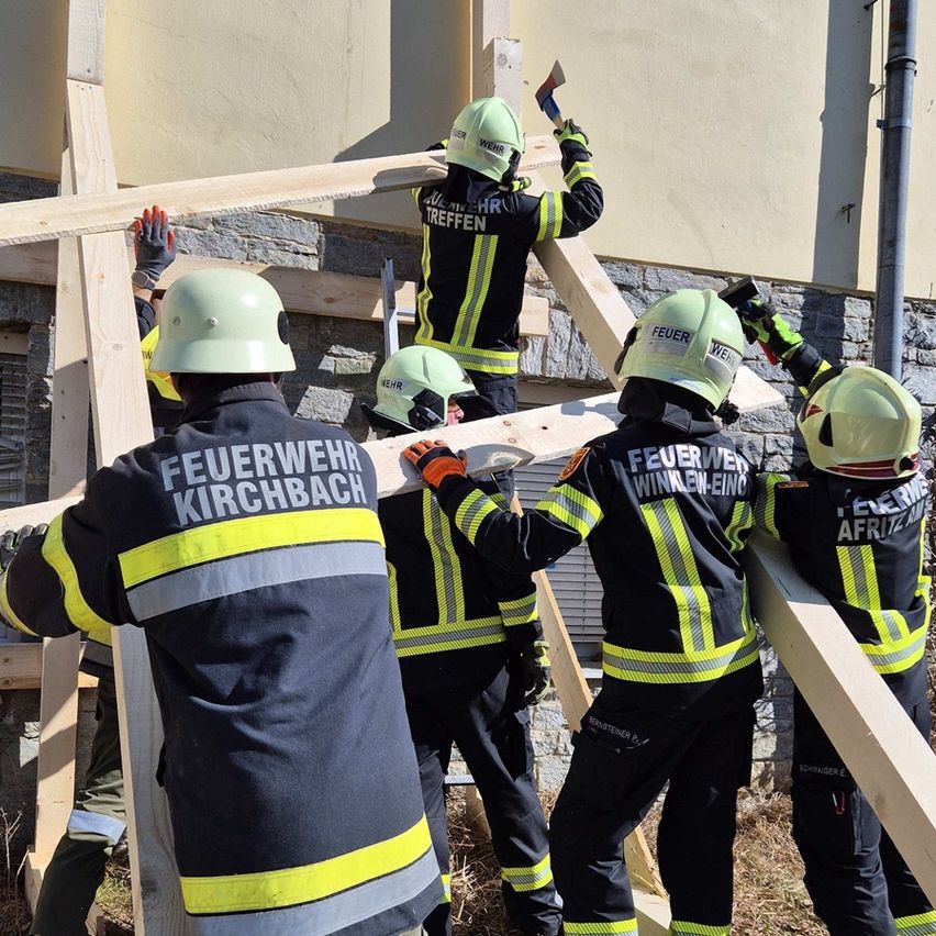 Eine Gruppe von Feuerwehrleuten, die Helme und Uniformen tragen, arbeiten zusammen, um eine Struktur mit Holzbrettern zu bauen. Sie sind draußen in der Nähe eines Gebäudes.