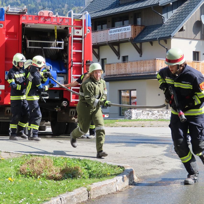 Feuerwehrleute in Uniformen ziehen einen Schlauch von einem LKW, während einer läuft und andere helfen. Ein Gebäude mit Balkonen und einem Schild, das 'NEUE WOHNUNGEN ZUM VERKAUF' anzeigt, befindet sich hinter ihnen.