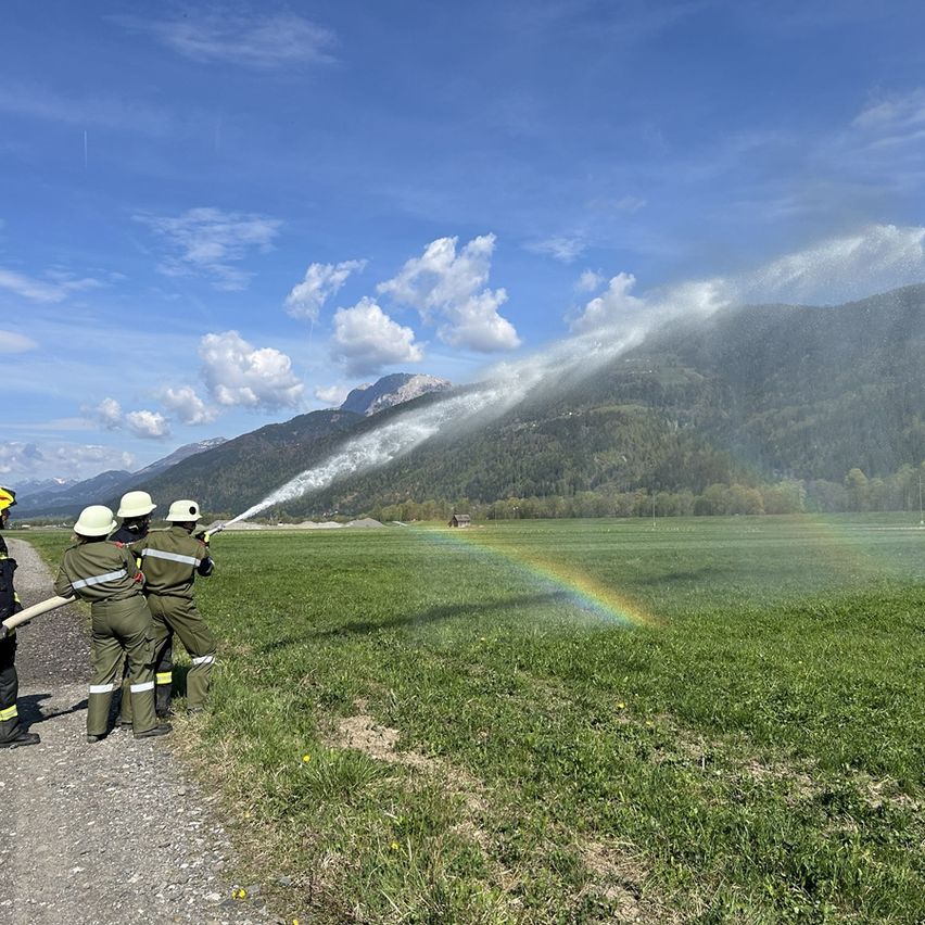 Drei Feuerwehrleute in Uniform sprühen Wasser aus einem Schlauch und erzeugen einen Regenbogen an einem sonnigen Tag mit Bergen im Hintergrund.