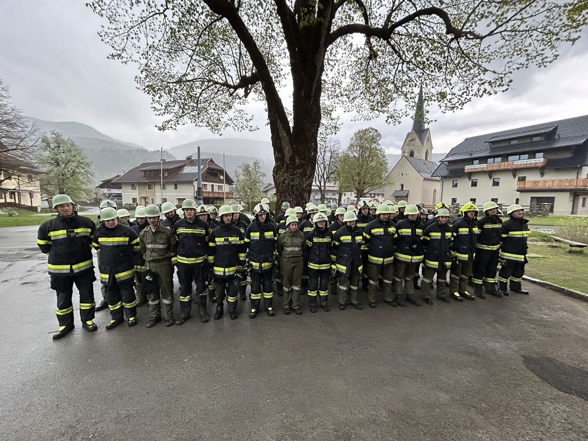 Eine Gruppe Feuerwehrleute in grünen Uniformen posiert unter einem Baum mit einer Kirche im Hintergrund.