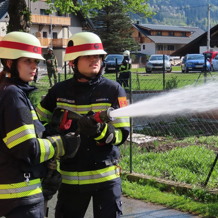 Zwei Feuerwehrleute in voller Ausrüstung üben mit einem Wasserschlauch im Freien, mit Gebäuden und geparkten Autos im Hintergrund.