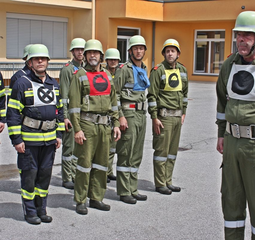 Eine Gruppe Feuerwehrleute in grünen Uniformen mit Helmen steht vor einem gelben Gebäude.