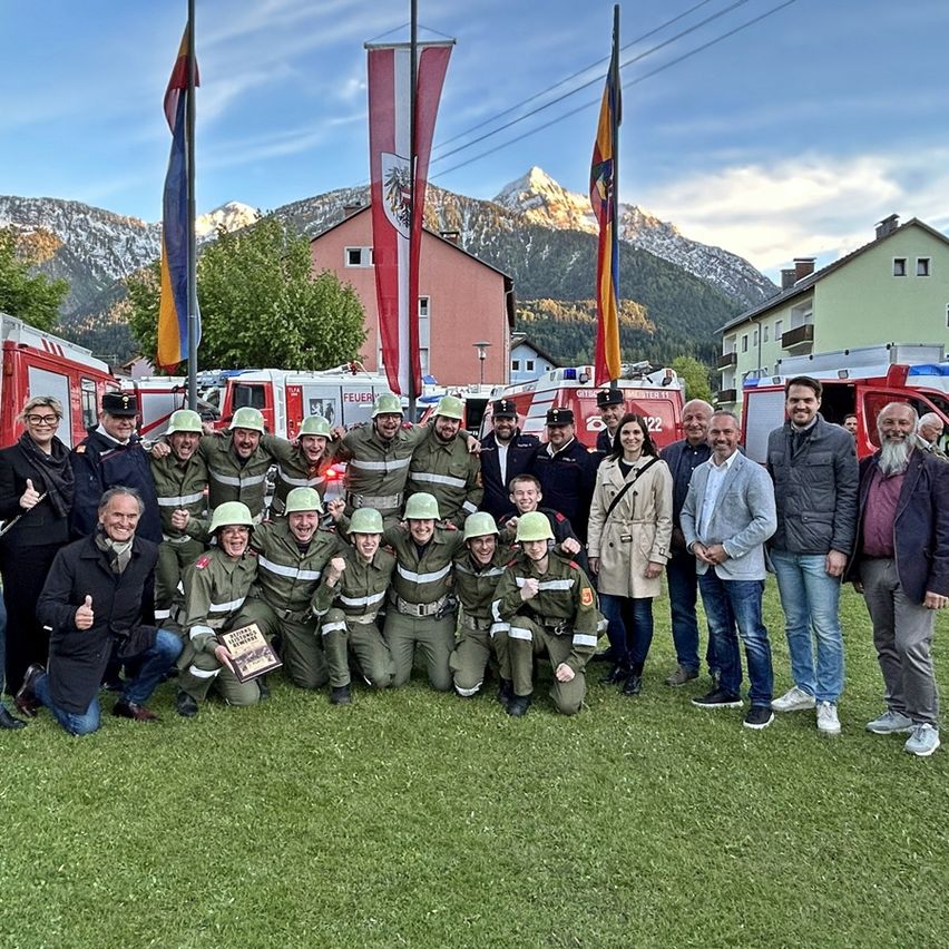 Eine Gruppe von Feuerwehrleuten posiert für ein Foto auf einem Rasen, mit Feuerwehrautos und Fahnen im Hintergrund. Sie alle lächeln und ein Mann hält einen Pokal.
