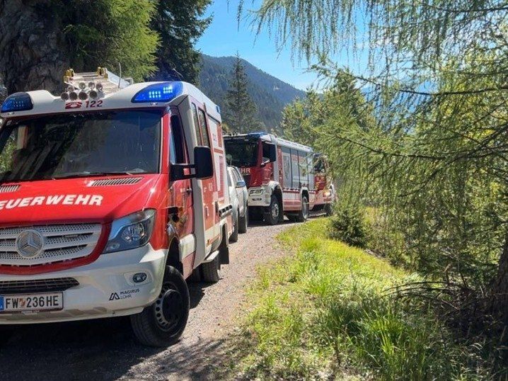 A red emergency vehicle with the number 122 is parked on a gravel road with other vehicles, surrounded by trees and grass. The sky is clear.