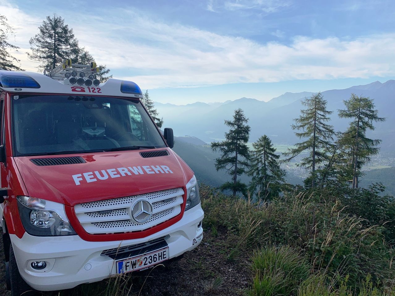 A red and white fire truck with the word Feuerwehr on the front, parked on a mountain road with a view of the mountains and sky.