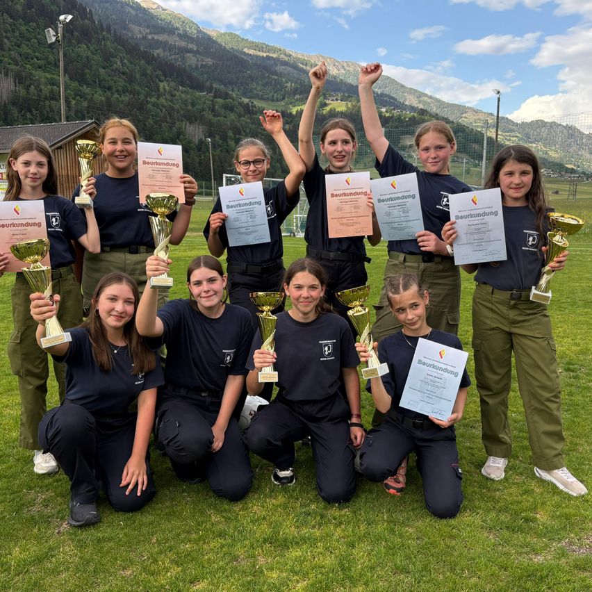 A group of young women in black uniforms pose for a photo on a green field with mountains in the background. They hold trophies and certificates.