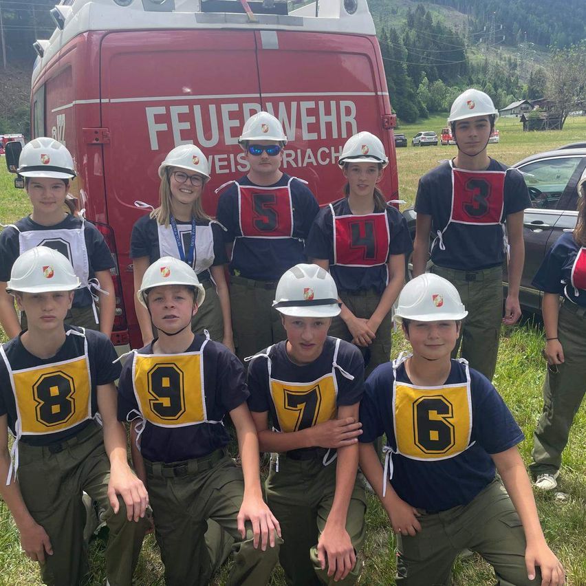 A group of young people wearing white helmets and numbered uniforms pose for a photo in front of a red fire truck.