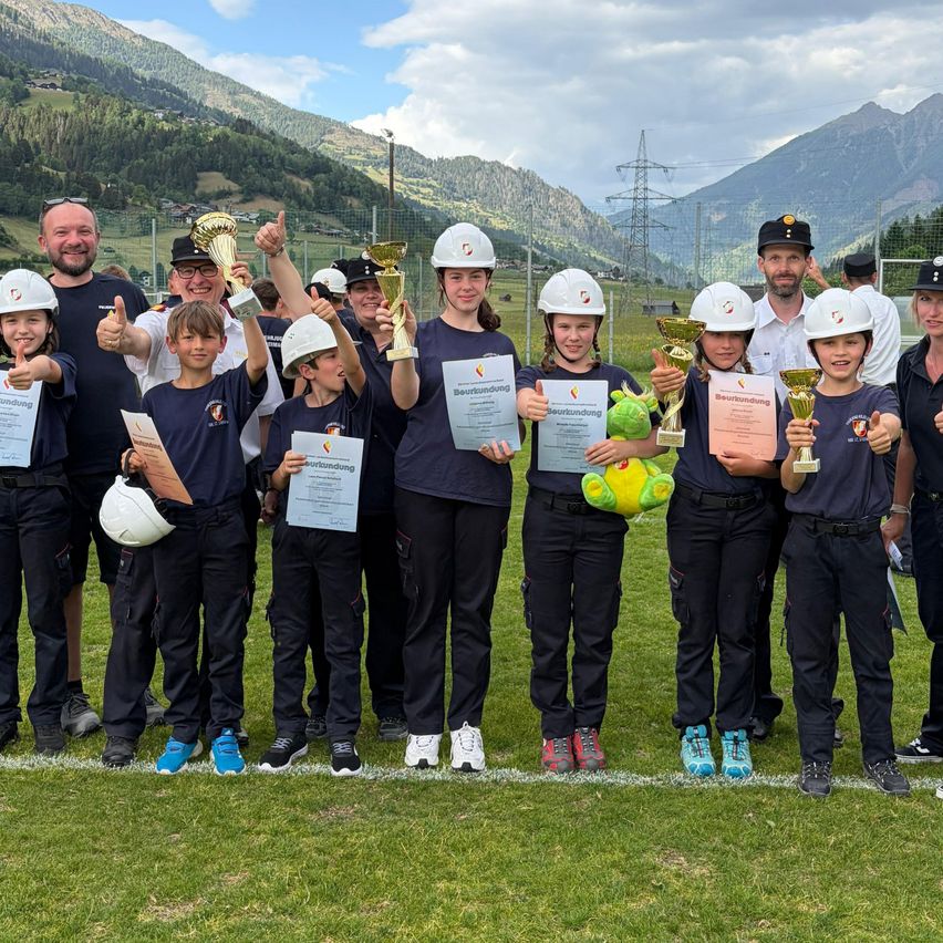 A group of children and adults are standing on a grassy field with mountains in the background. They are wearing uniforms and holding certificates and trophies.