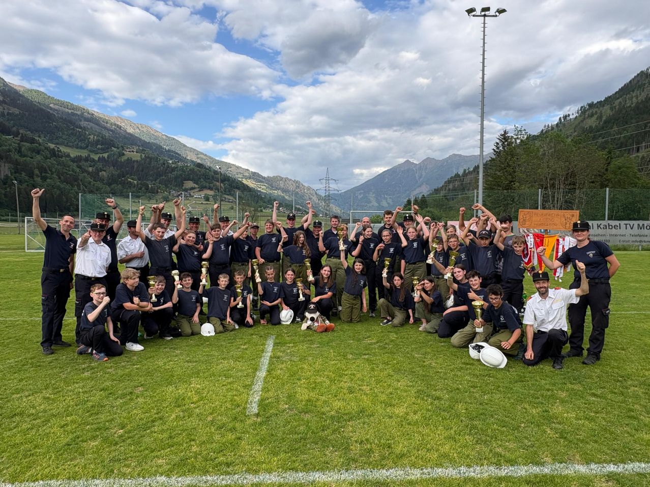 A group of individuals dressed in blue uniforms are celebrating on a sports field with mountains in the background. They hold trophies and are posing for a photo.