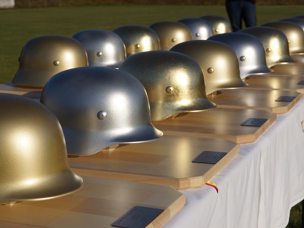 A row of golden and silver helmets are on a table. They are lined up, with some of them having tags. The background is blurry, with a green field and a person standing.