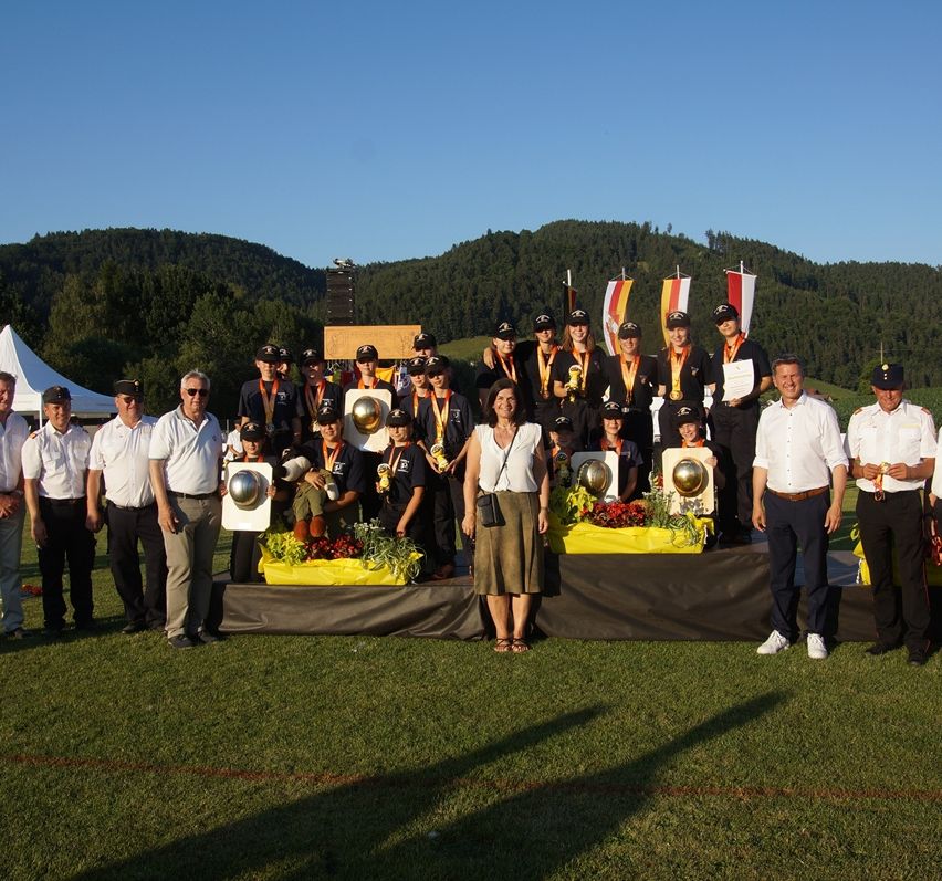 A group of people on a field, some holding medals, stand behind a stage with trophies, flowers, and flags. Behind them, there are tents and mountains.