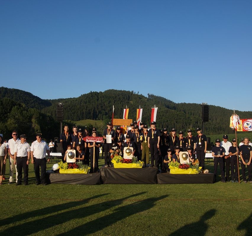 A group of people wearing medals stand on a stage with flowers in front of a crowd. The stage has a black backdrop. Behind them are mountains and flags. Some of them are holding trophies.