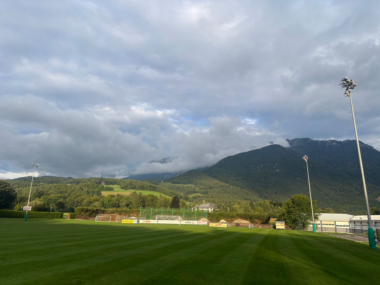 Ein leerer Fußballplatz mit grünem Rasen und bewölktem Himmel. Im Hintergrund befindet sich ein großes Bergmassiv und ein Fußballtor auf der linken Seite.