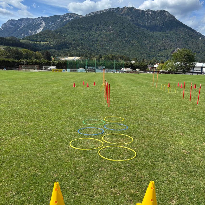 Ein Rasenfeld mit Bergkulisse, das eine Reihe bunter Hula-Hoops und in einem Zickzack-Muster angeordneter Kegel aufweist, wobei im Hintergrund Fußballtore zu sehen sind.