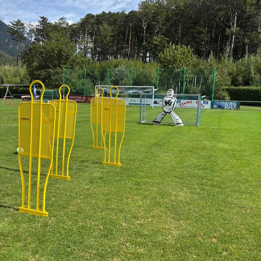 Ein Fußballfeld mit gelben Torpfosten und einer großen aufblasbaren Torwartfigur vor dem Tor. Das Feld ist von Bäumen und Büschen umgeben.