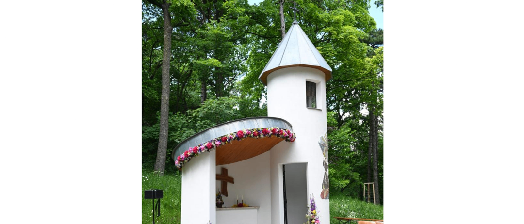Eine kleine Kapelle mit einem Kreuz auf dem Dach, umgeben von Blumen und Grün. Ein Tisch mit einem weißen Tuch und einem Kreuz darauf. Ein Lautsprecher auf einem Stativ ist in der Nähe.