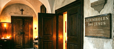 Eine schwach beleuchtete Kapelle mit Holztüren und Bänken. Ein Kreuz ist an der Wand über der Tür angebracht. Der Text an der Wand lautet 'Atemho bei Jesus'.