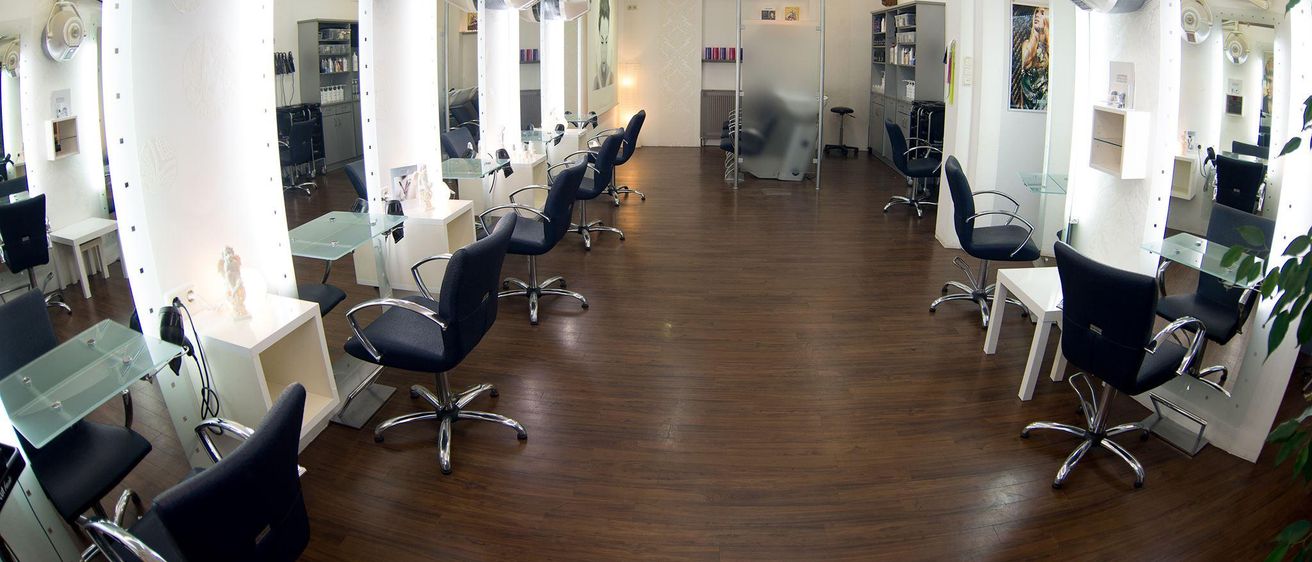Ein leerer Salon mit Holzböden, Glastischen, schwarzen Drehstühlen und Deckenleuchten. Eine Person ist im Hintergrund verschwommen.