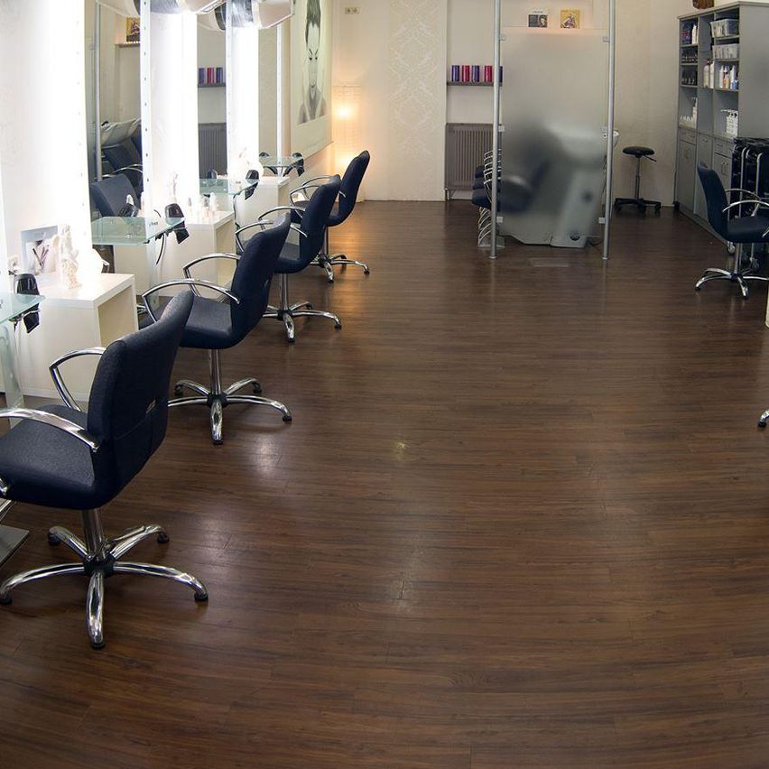 Ein leerer Salon mit Holzböden, Glastischen, schwarzen Drehstühlen und Deckenleuchten. Eine Person ist im Hintergrund verschwommen.