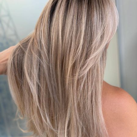 Eine Nahaufnahme einer Frau mit langen, hellbraunen Haaren, die in sanften Wellen gestylt sind, von hinten betrachtet, mit einem unscharfen Hintergrund.