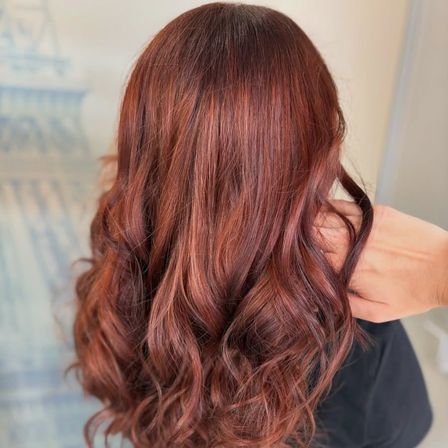 Eine Frau mit langen, welligen, rotbraunen Haaren hält ihr Haar mit einer Hand, möglicherweise drinnen.