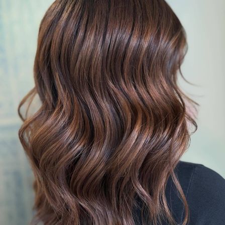 Nahaufnahme des Rückens einer Frau mit schulterlangem, gewelltem braunem Haar vor hellem Hintergrund.