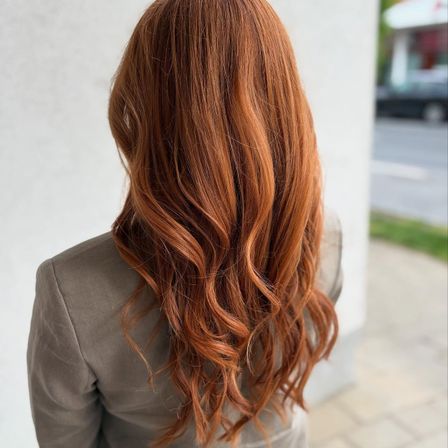 Eine Frau mit langen, welligen roten Haaren steht mit dem Rücken zugewandt und trägt einen grauen Blazer. Sie befindet sich draußen in der Nähe einer weißen Wand, mit einem verschwommenen Auto und einem Grasbereich im Hintergrund.
