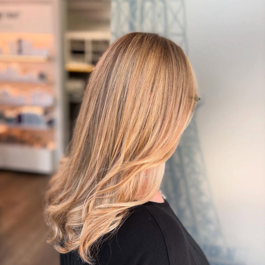 Rückansicht einer Frau mit langen, welligen blonden Haaren, die eine Brille und ein schwarzes Oberteil trägt, in einem Salon.