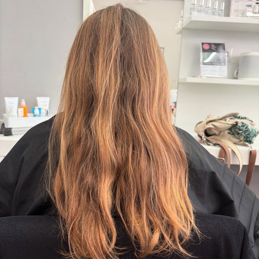 Eine Frau mit langen, blonden Haaren sitzt in einem Salon und trägt einen schwarzen Umhang. Hinter ihr befindet sich ein weißer Regal mit verschiedenen Gegenständen.