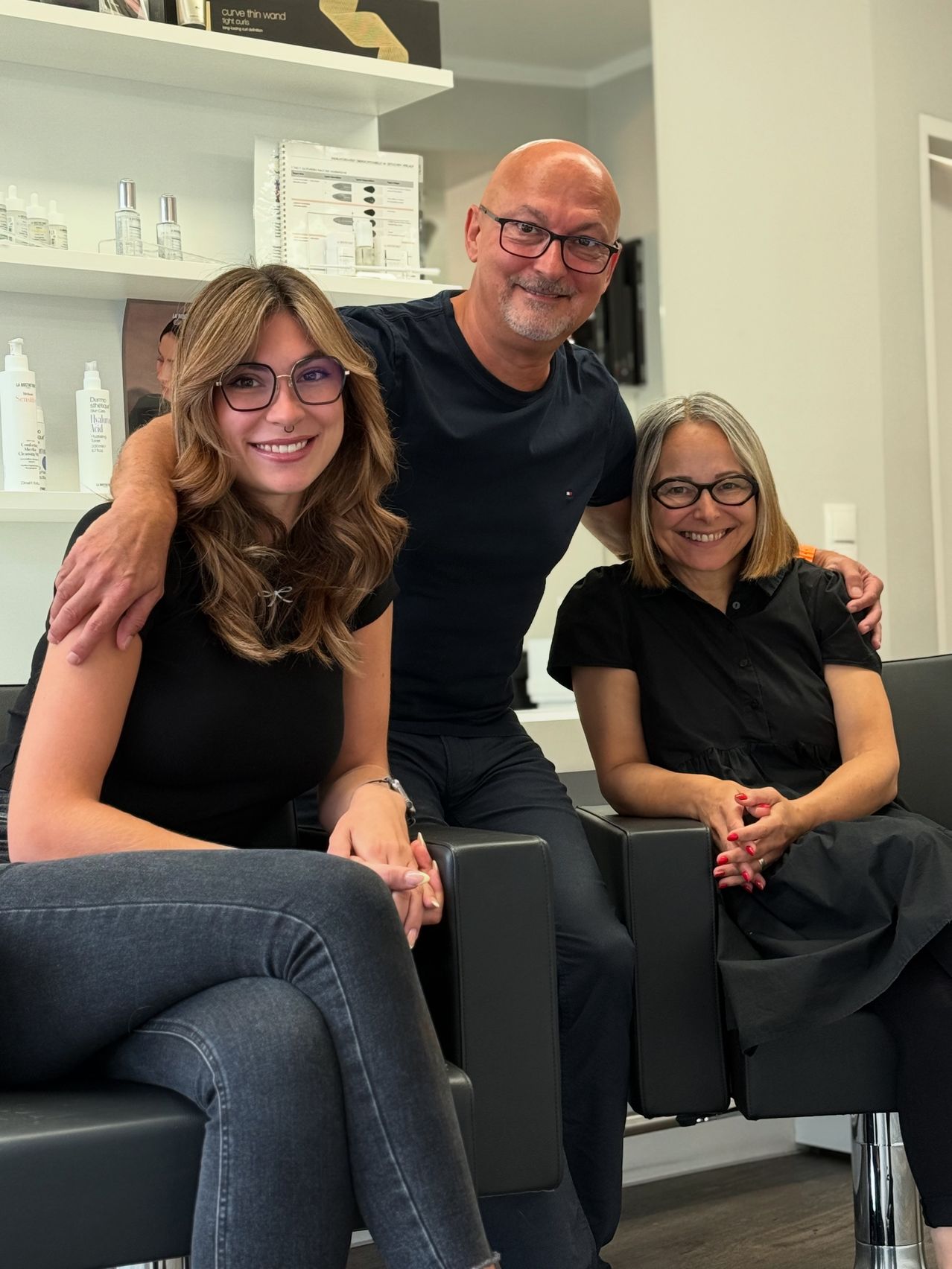 Drei Menschen sitzen auf schwarzen Sesseln in einem Salon. Zwei Frauen und ein Mann. Der Mann hat seinen Arm um die Frau auf seiner linken Seite gelegt.