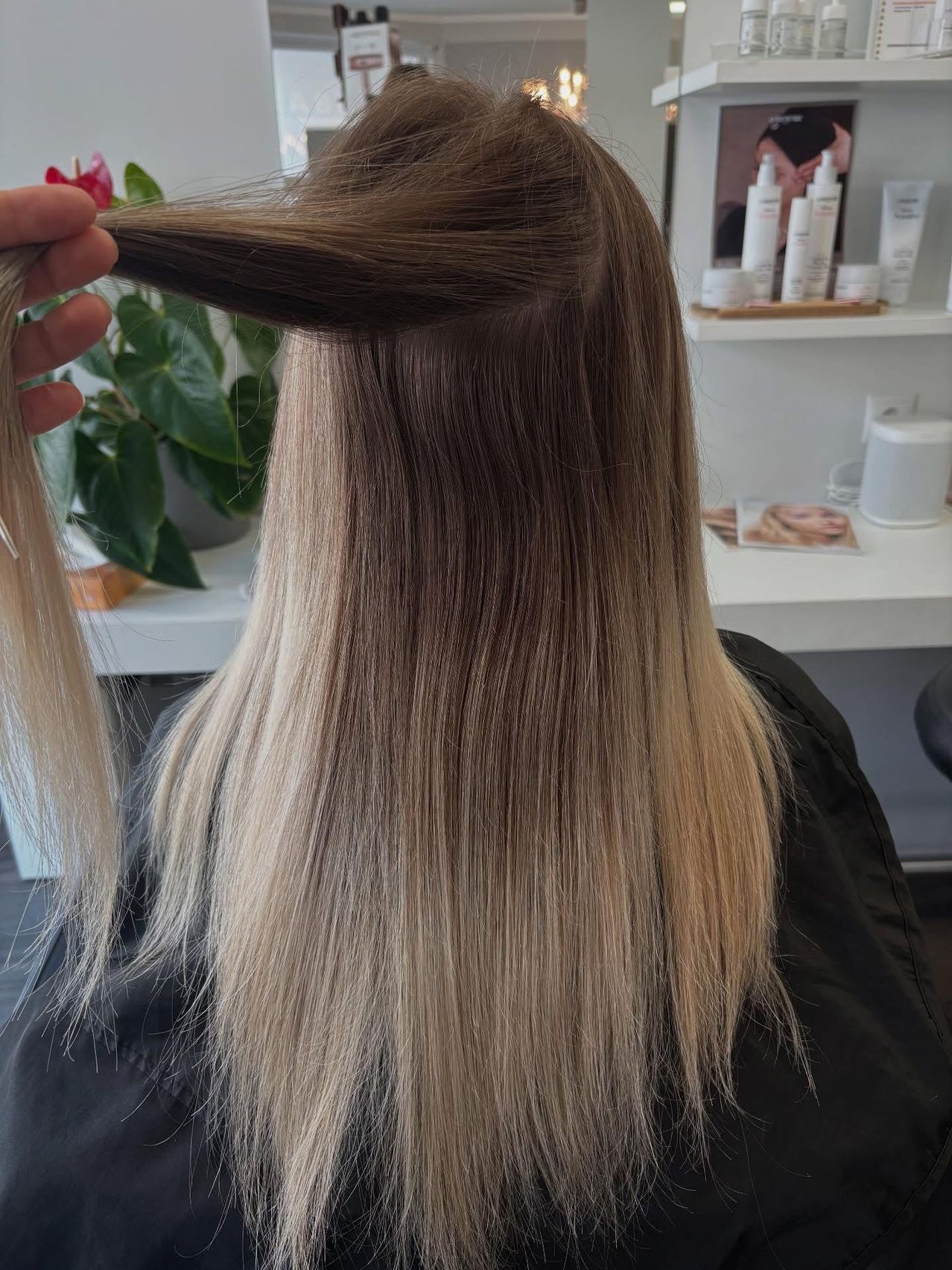 Eine Person hält eine Strähne blonder Haare, wahrscheinlich in einem Salon. Die Haare sind lang und haben einen Farbverlauf von hell bis dunkel. Dahinter steht ein weißer Regal mit Haarpflegeprodukten.