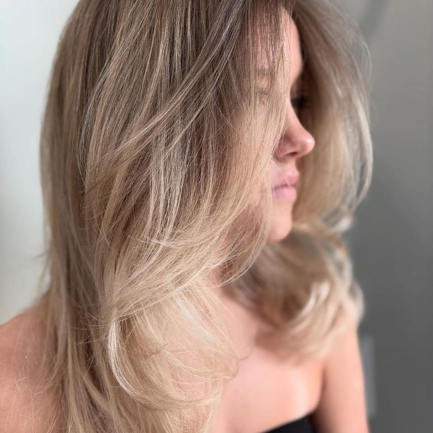 Nahaufnahme einer Frau mit langen, welligen blonden Haaren, ihr Gesicht ist zur Seite gewandt. Sie hat ein sanftes Lächeln und trägt ein schwarzes ärmelloses Oberteil.