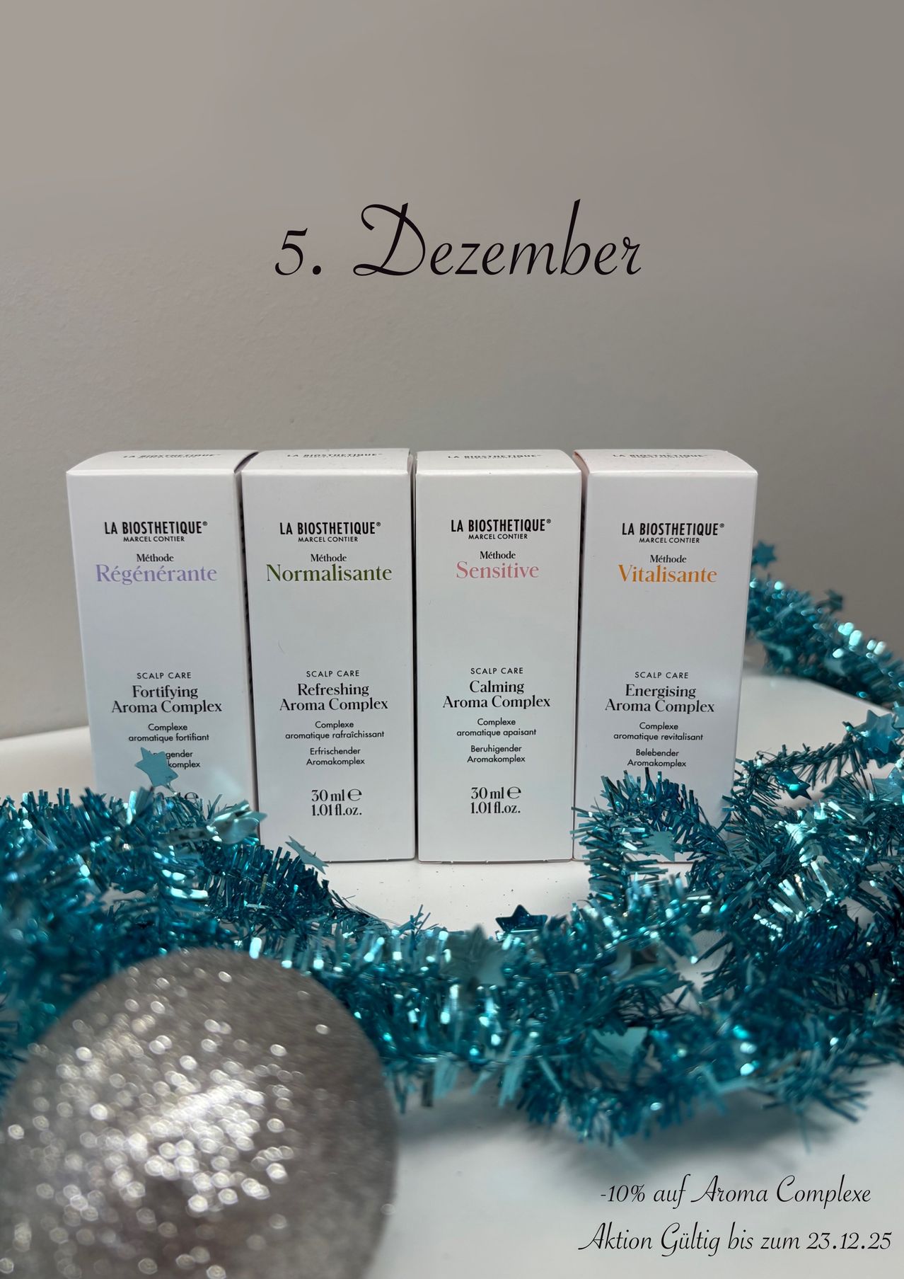 Fünf Schachteln mit Kopfhautpflegeprodukten von La Biosthetique mit Etiketten wie Regenerierend, Normalisierend, Empfindlich und Vitalisierend sind auf einer weißen Oberfläche mit blauem Tinsel angeordnet. Der 5. Dezember ist in Lila auf einer weißen Wand geschrieben.