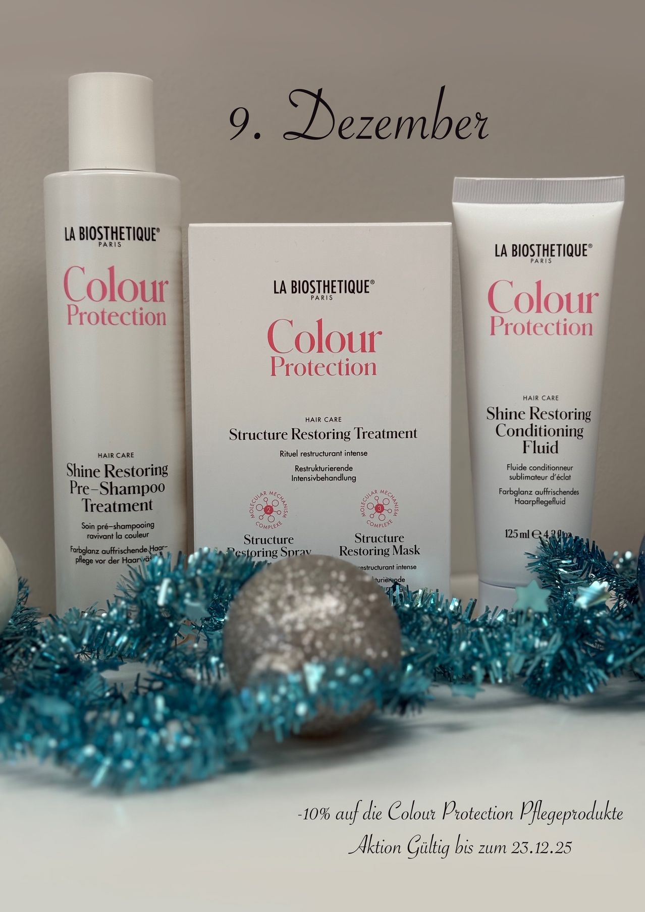 Drei Flaschen La Biosthetique Colour Protection Haarpflegeprodukte stehen auf einer weißen Oberfläche, umgeben von blauer Tinsel und einer silbernen Kugel. Die Produkte umfassen ein Shine Restoring Pre-Shampoo Treatment, ein Structure Restoring Treatment und ein Shine Restoring Conditioning Fluid.