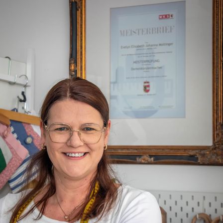 Eine Frau in einem weißen Shirt und Brille lächelt vor einem gerahmten Zertifikat und mehreren Kleidungsstücken.